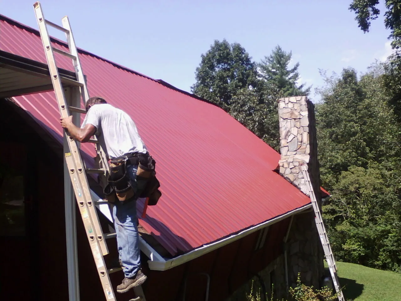 Roofer on a ladder beside a red metal roof and stone chimney — Soffit & Fascia Repair by Roofing Brothers in Sprague